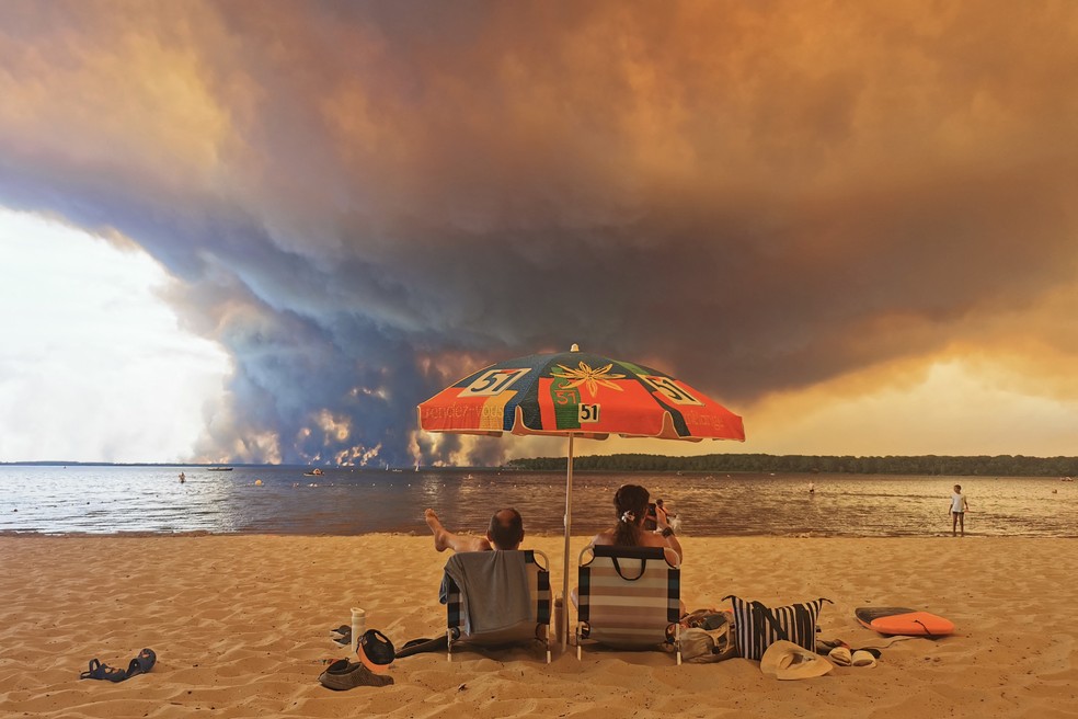 Banhistas à beira do lago Lacanau, no sudeste da França, com incêndio florestal ao fundo na sexta (24) — Foto: MAXIMILIEN LAMY / AFP
