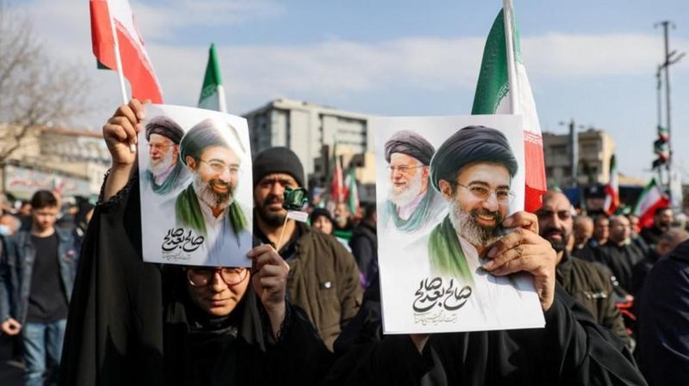 Apoiadores mostram cartazes com Mojtaba Khamenei ao lado do seu pai, o aiatolá Ali Khamenei (1939-2026), em manifestações realizadas em Teerã — Foto: WANA via REUTERS via BBC