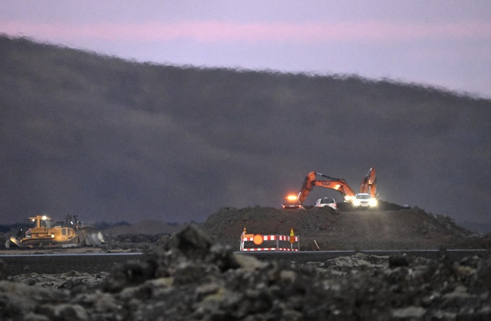 Dois tratores trabalham numa barreira de proteção para evitar que o fluxo de lava chegue ao centro da cidade de Grindavik, no sudoeste da Islândia, após uma erupção vulcânica neste domingo, 14 de janeiro de 2024. — Foto: Halldor KOLBEINS/AFP
