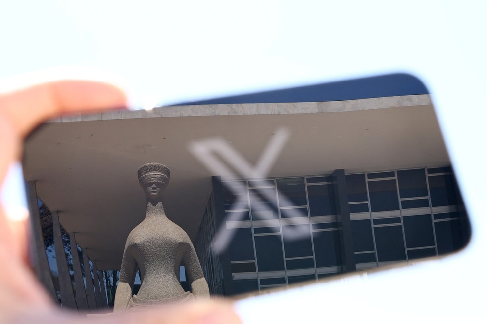 La estatua de la Justicia, ubicada frente al edificio del Supremo Tribunal Federal de Brasil, reflejada en la pantalla de un teléfono móvil que exhibe el logotipo de la red social X, anteriormente conocida como Twitter.