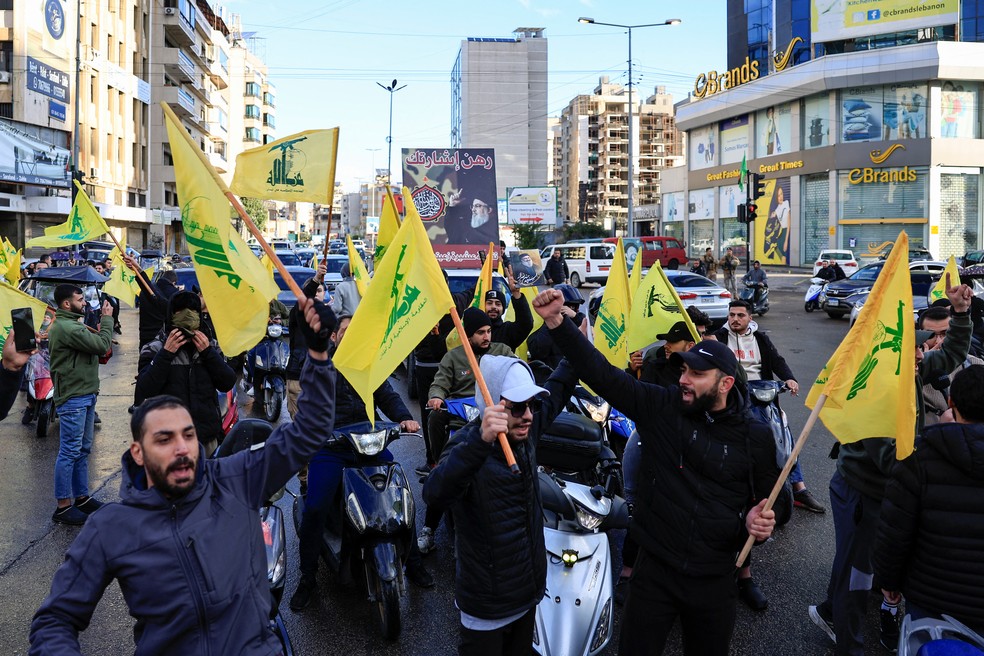 Homens fazem gestos enquanto seguram bandeiras do Hezbollah, na entrada dos subúrbios do sul de Beirute — Foto: Thaier Al-Sudani/Reuters