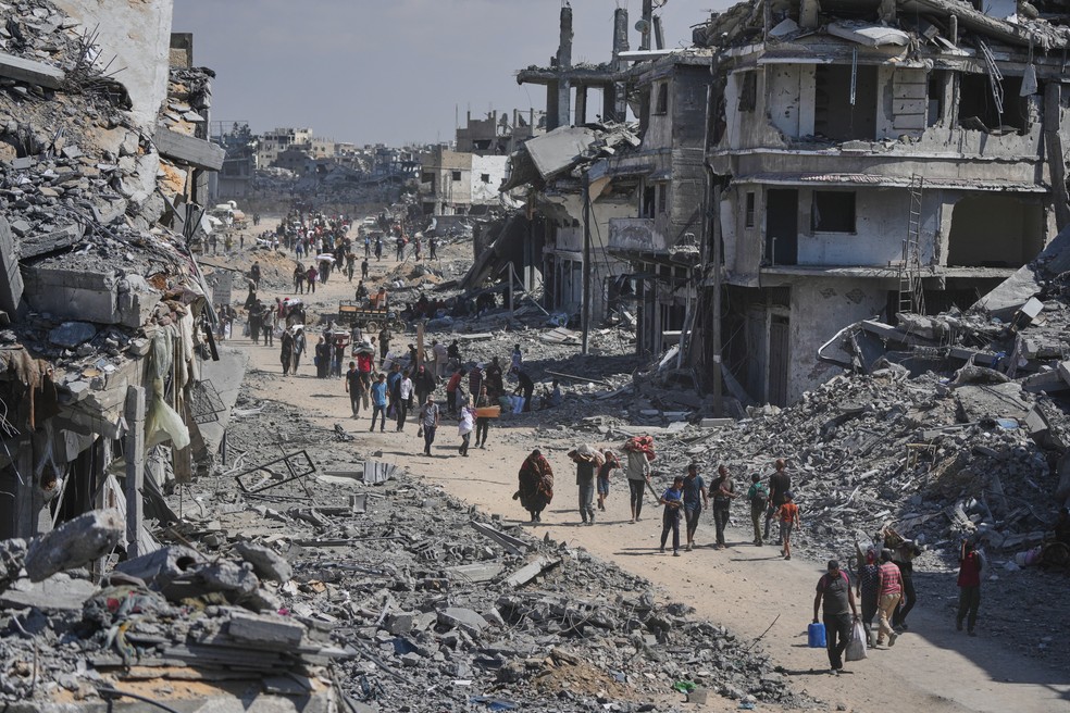 Palestinos deslocados caminham com seus pertences em frente a prédios destruídos enquanto retornam para suas casas em Khan Younis, no sul da Faixa de Gaza, sexta-feira, 10 de outubro de 2025 — Foto: AP Photo/Jehad Alshrafi