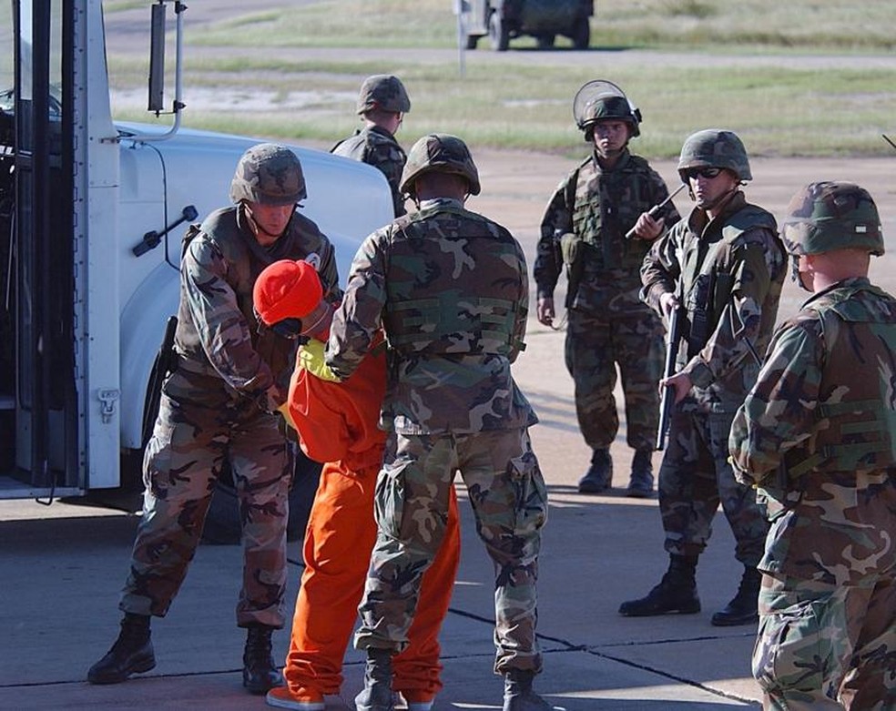As imagens mostram a grande expectativa ao redor da chegada dos primeiros prisioneiros a Guantánamo. A transferência dos prisioneiros do avião para o ônibus que os transportaria para a prisão foi fortemente vigiada por fuzileiros navais prontos para agir. — Foto: JEREMY T. LOCK/USAF via BBC
