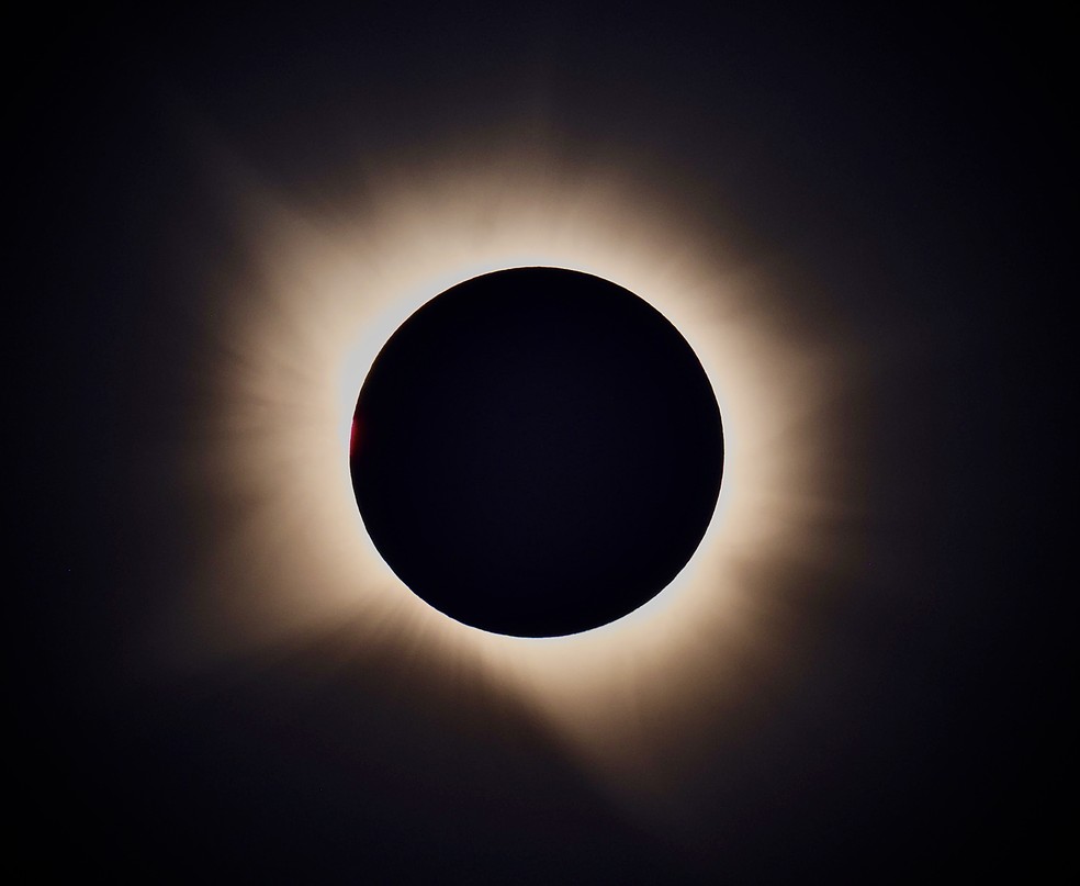Proeminências solares aparecem em tons de rosa e vermelho ao redor do disco encoberto pela Lua. As estruturas de plasma se estendem para além da superfície visível do Sol. — Foto: Kiko Fairbairn / @kikofairbairn