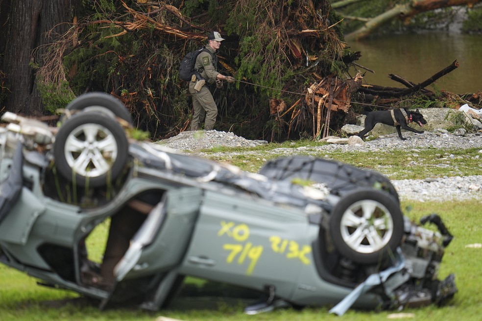 Policial faz buscas com cachorro (ao fundo) por desaparecidos diante de carro revirado por causa das enchentes após chuvas no Texas, em 5 de julho de 2025. — Foto: Julio Cortez/ AP