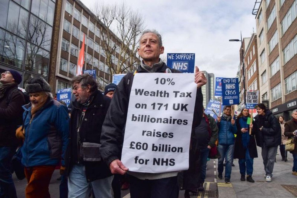O ativista Peter Tatchell segura um cartaz pedindo imposto sobre a riqueza dos bilionários para financiar o sistema de saúde britânico em 2023 em Londres — Foto: GETTY IMAGES via BBC