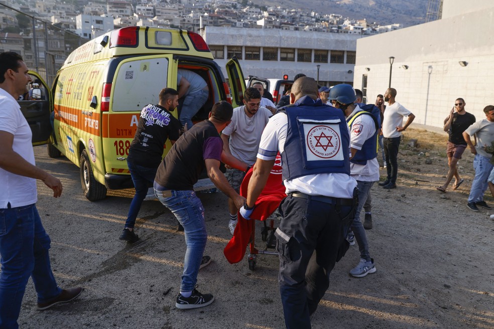 Feridos são resgatados nas Colinas de Golã, região controlada por Israel, após um foguete atingir um campo de futebol da aldeia local — Foto: Forças de segurança e médicos israelitas transportam feridos, juntamente com residentes locais, no local onde caiu um ataque do Líbano, na aldeia de Majdal Shams, na zona de Golã anexada a Israel, a 27 de julho de 2024 - Foto: Jalaa MAREY / AFP