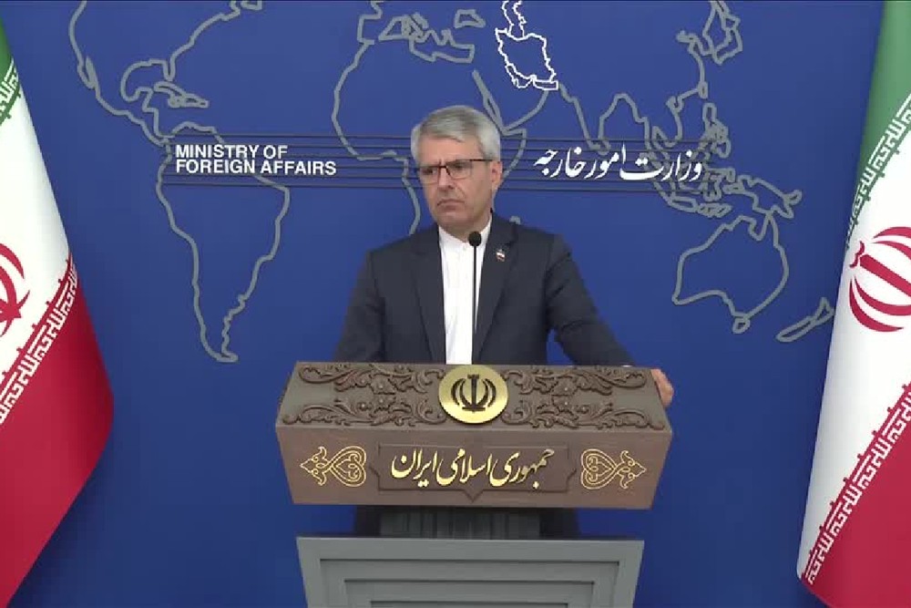 Coletiva de imprensa com porta-voz em frente a um mapa-múndi ao fundo e ao púlpito com o símbolo do governo iraniano; três bandeiras aparecem nas laterais.