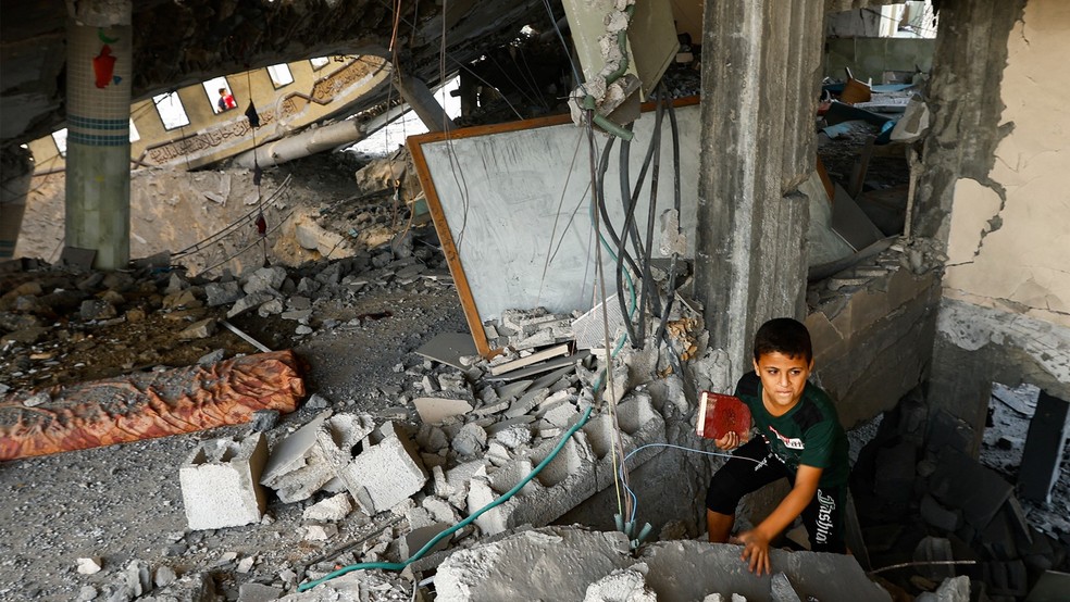 Um menino carrega um livro enquanto palestinos inspecionam uma mesquita destruída pelos ataque — Foto: Reuters