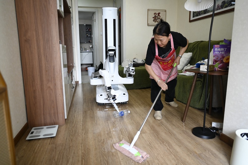 Foto tirada em 21 de maio de 2026, mostra uma empregada doméstica trabalhando ao lado de um robô da X Square na casa de um cliente em Pequim — Foto: WANG ZHAO/AFP