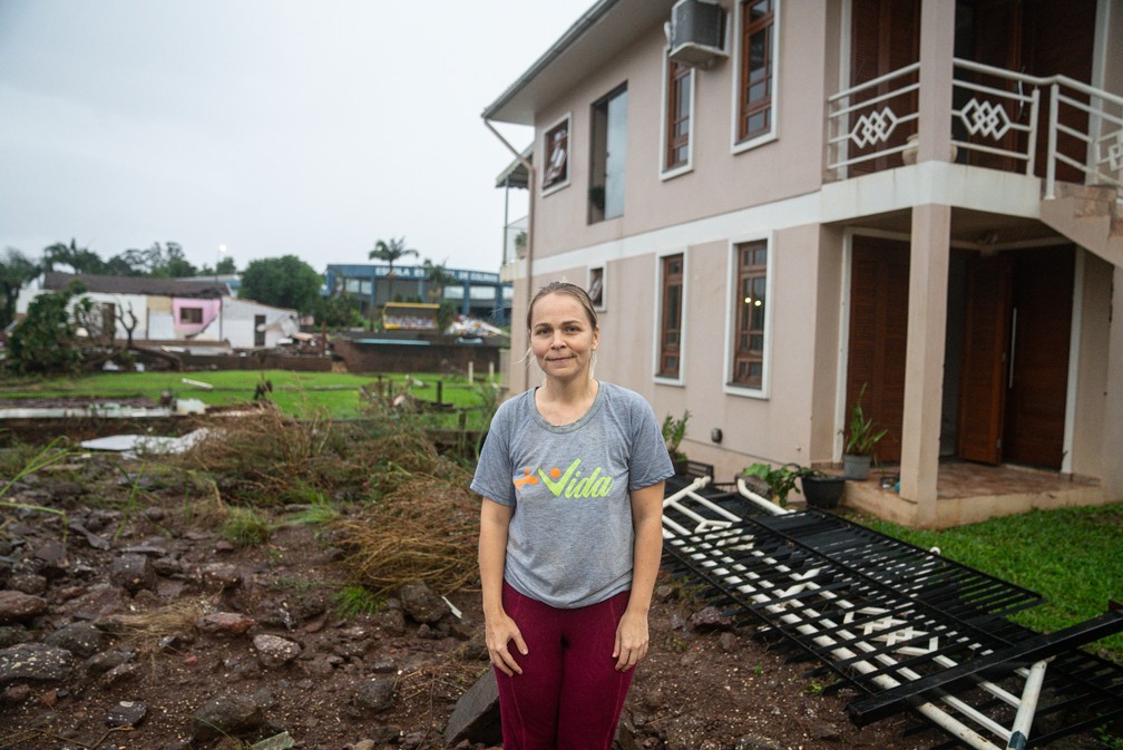 Patrícia Knobloch posa ao lado de sua casa em Colinas (RS). Ao fundo, a casa da máe dela, que foi destruída pela enchente — Foto: Fábio Tito/g1