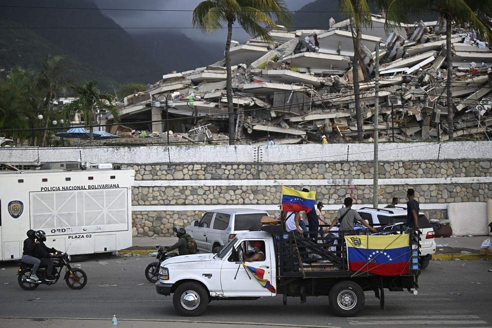 Um caminhão com bandeiras venezuelanas passa por um edifício desabado após dois terremotos, em Los Corales, no estado de La Guaira, a cerca de 40 km ao nordeste de Caracas, em 27 de junho de 2026. — Foto: FEDERICO PARRA/AFP