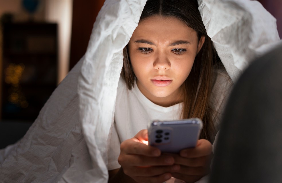 Adolescente na cama mexendo no celular. — Foto: Freepik