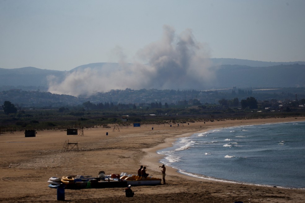 Pessoas na praia e, ao fundo, a fumaça após ataques na região sul do Líbano nesta quinta (26) — Foto: Amr Abdallah Dalsh/Reuters