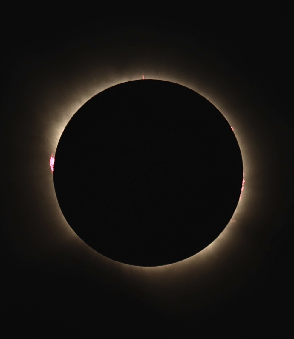 A coroa solar aparece como um grande halo branco ao redor da Lua durante o eclipse total. À esquerda, uma proeminência solar forma uma pequena região avermelhada. — Foto: Kiko Fairbairn / @kikofairbairn