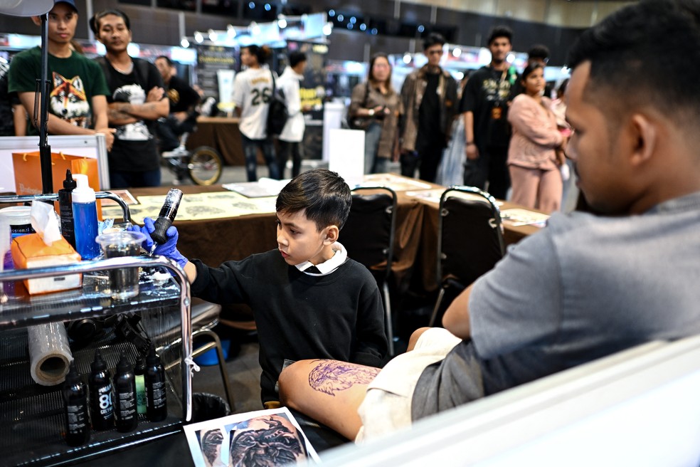 Visitantes de feira de tatuagem na Tailândia, observam Napat Mitmakorn, tatuador de 9 anos, em março de 2025. — Foto: Manan Vatsyayana/ AFP