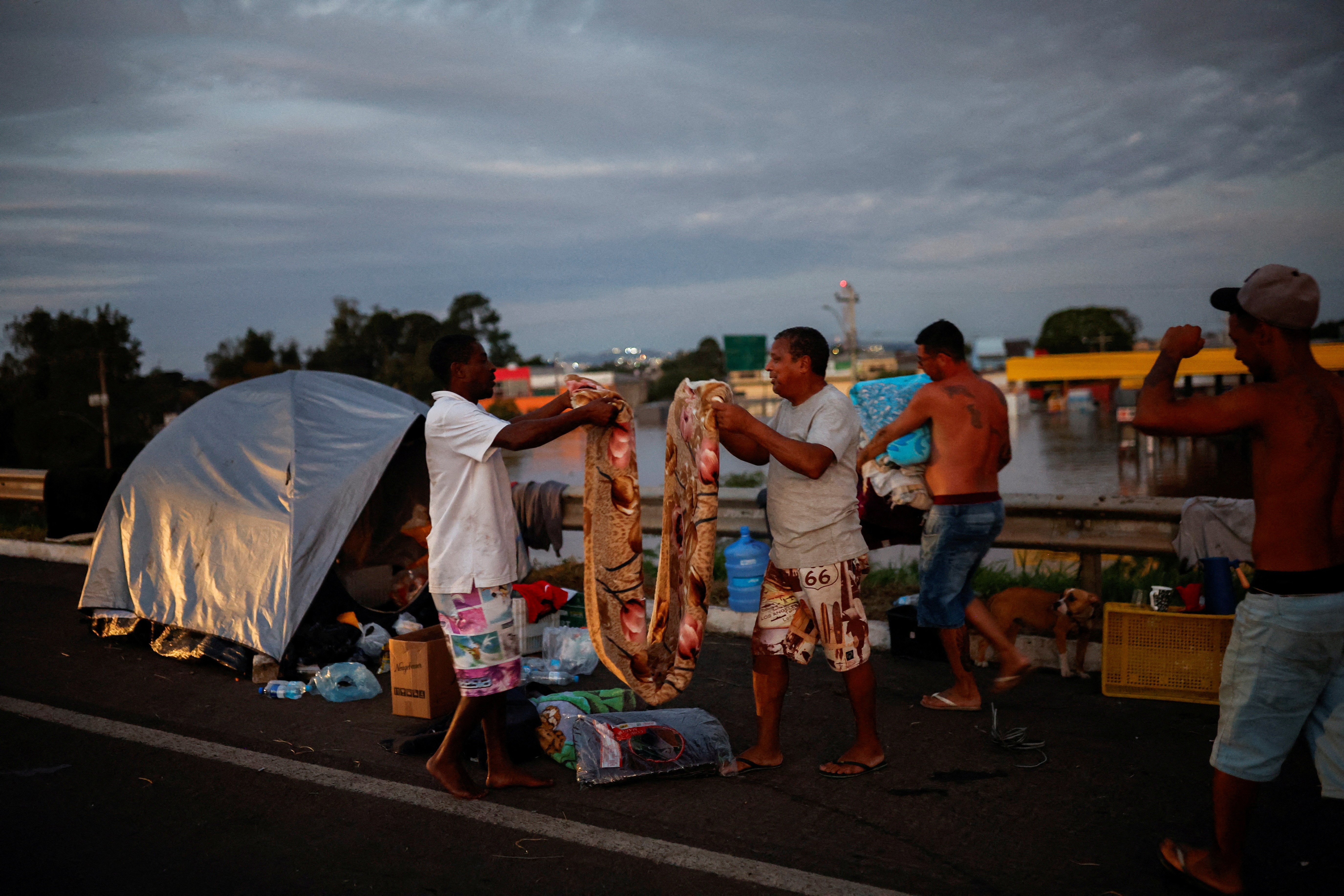 Homens dobram um cobertor enquanto acampam na estrada perto de uma rua inundada em Eldorado do Sul, no Rio Grande do Sul, Brasil, 6 de maio de 2024.