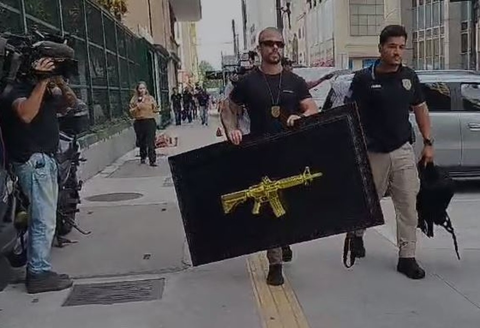 Policial carrega quadro com réplica de fuzil dourado na residência de 'Cigarreiro' — Foto: Reprodução