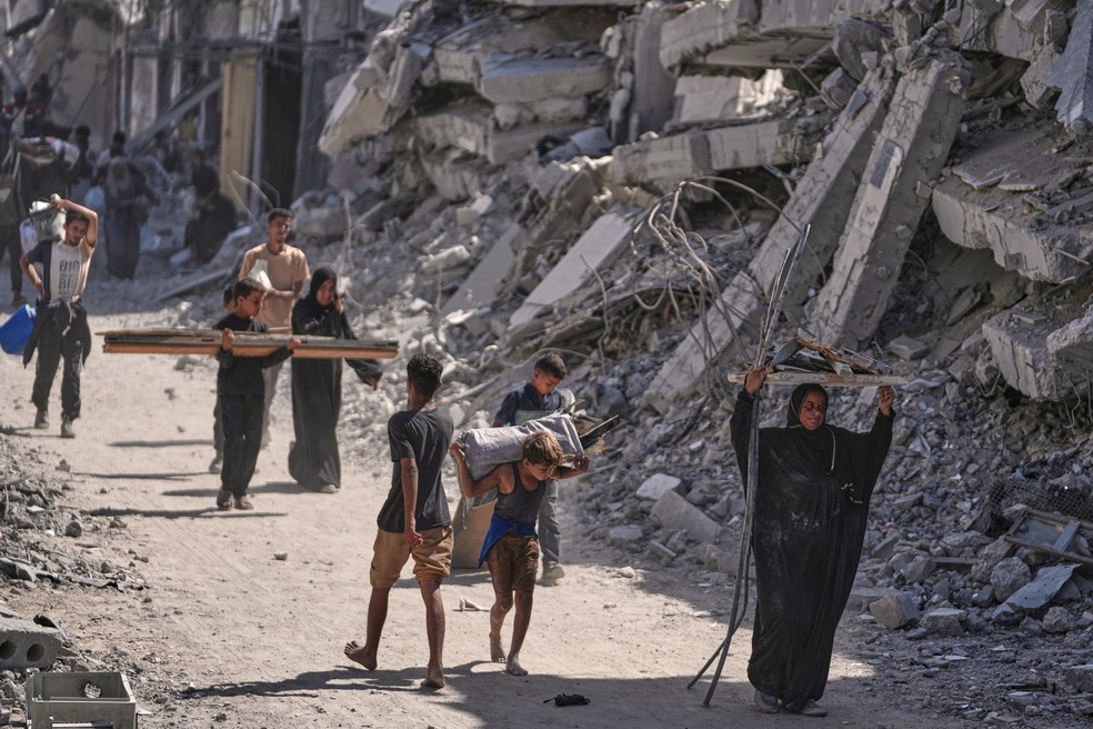 Palestinos deslocados caminham com seus pertences em frente a prédios destruídos enquanto retornam para suas casas em Khan Younis, no sul da Faixa de Gaza — Foto: AP Photo/Jehad Alshrafi