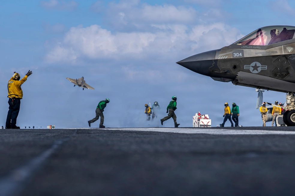 Jato F-35 (à direita) estacionado no deque do porta-aviões USS Abraham Lincoln enquanto jato F-18 decola (ao fundo) para voo de monitoramento em 23 de janeiro de 2026. — Foto: Daniel Kimmelman/Marinha dos Estados Unidos