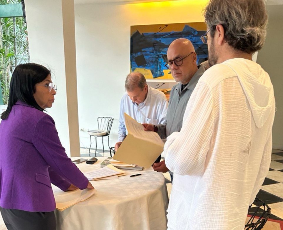 Edmundo González (ao fundo) na embaixada da Espanha em Caracasm, acompanhado da vice-presidente da Venezuela, Delcy Rodríguez (à esq), e do presidente da Assembleia Nacional, Jorge Rodríguez (careca), assinando documento reconhecendo vitória de Maduro na eleição. Assinatura ocorreu no início de setembro. — Foto: Reprodução/Jorge Rodríguez