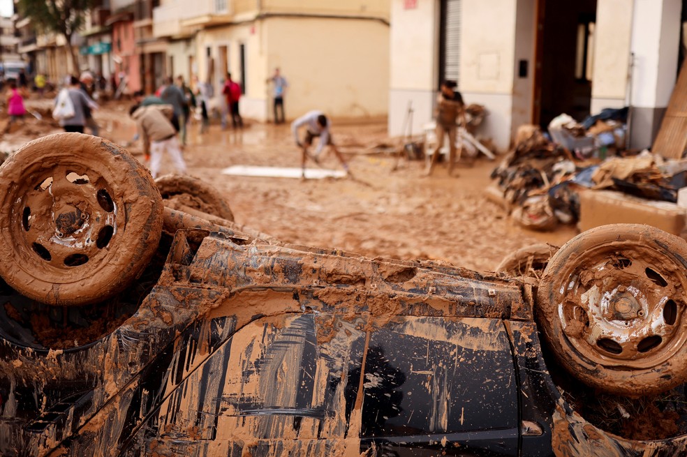 Enchentes na Espanha: veículo danificado é visto em Paiporta, perto de Valência. Ao fundo, voluntários trabalham na limpeza da lama. — Foto: REUTERS/Nacho Doce