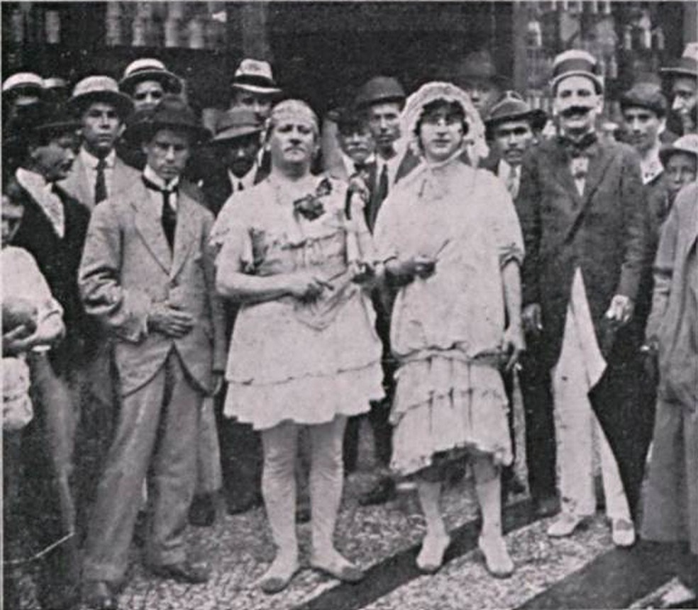 Homens de paletó ao lado de outros foliões fantasiados na Avenida Rio Branco, entre eles vestidos de mulher em foto de 1916 do jornal 'Careta' — Foto: Acervo da Fundação Biblioteca Nacional – Brasil