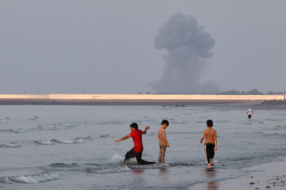 Três meninos brincam nas águas rasas do Estreito de Ormuz, enquanto uma coluna de fumaça sobe de uma explosão ao fundo, perto de Bandar Abbas, Irã — Foto: Razieh Poudat/ISNA via AP