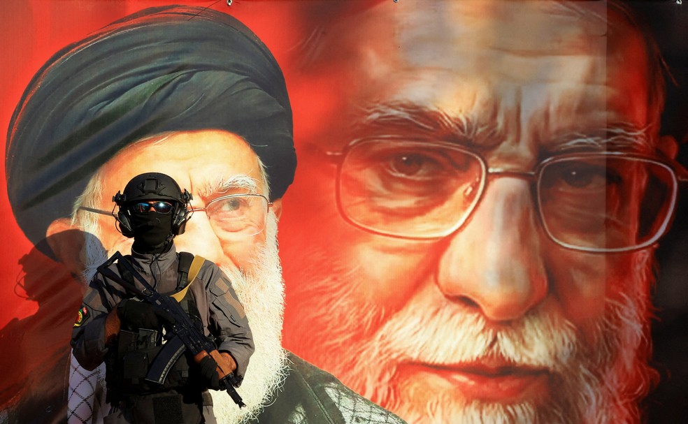 Um membro do Kataib Sayyid al-Shuhada faz guarda em frente a um mural que retrata o falecido Líder Supremo do Irã, o Aiatolá Ali Khamenei — Foto: REUTERS/Thaier al-Sudani
