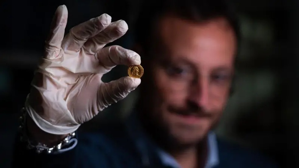 O arqueólogo Alessandro Russo segura uma moeda de ouro encontrada com o esqueleto feminino. — Foto: TONY JOLLIFFE/BBC