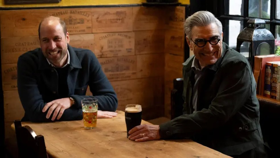 Príncipe William e Eugene Levy sorriem enquanto bebem uma Guinness e uma cidra em um pub próximo. — Foto: AppleTV+/PA Media via BBC