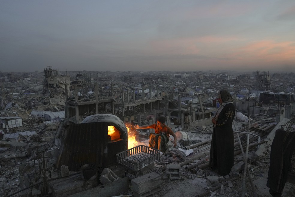 Menino cozinha ao lado da mãe no teto de uma casa destruída, na Faixa de Gaza, em 17 de março de 2025 — Foto: AP Photo/Jehad Alshrafi