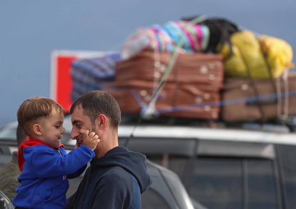 Refugiado na região de Nagorno-Karabakh segura criança pequena no colo diante de carro com malas amarradas no teto — Foto: Irakli Gedenidze/REUTERS