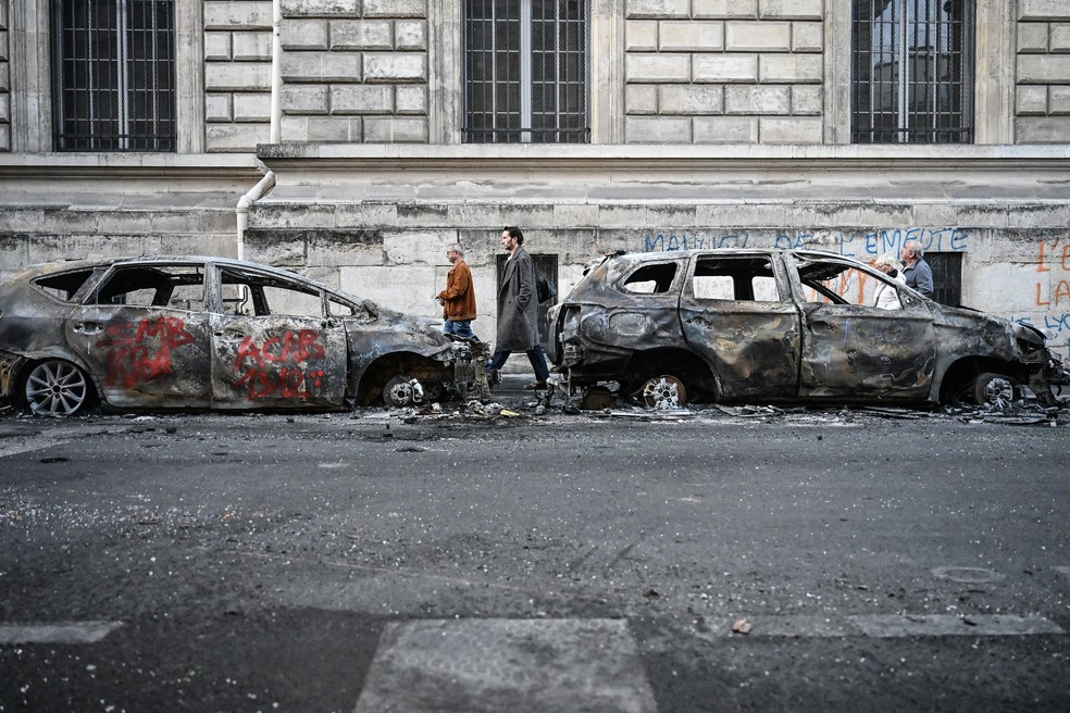 Pedestres passam por dois veículos queimados estacionados em uma rua um dia após os protestos nacionais contra o alto custo de vida, em Paris. — Foto: MATTHIEU RONDEL/AFP