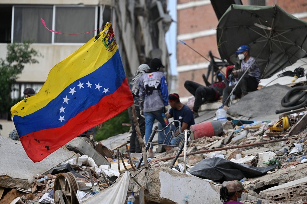 Uma bandeira venezuelana tremula em meio aos escombros de um prédio que desabou em Caraballeda, estado de La Guaira, Venezuela, em 9 de julho de 2026, após os terremotos de 24 de junho. — Foto: RAUL ARBOLEDA / AFP