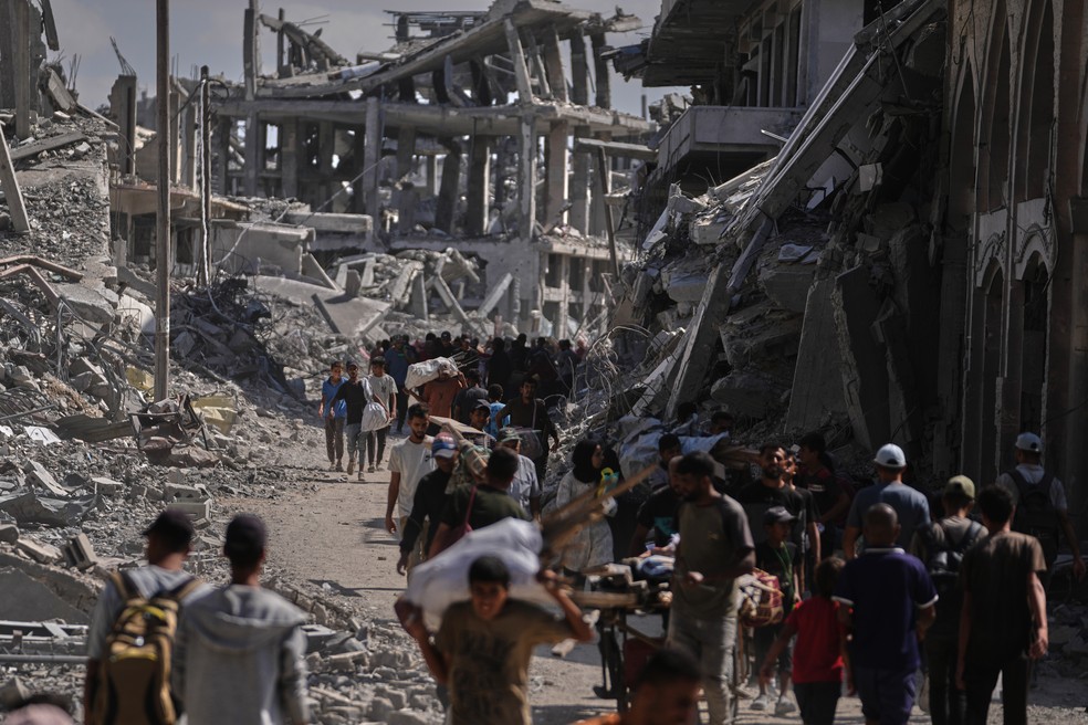 Palestinos deslocados caminham com seus pertences em frente a prédios destruídos enquanto retornam para suas casas em Khan Younis, no sul da Faixa de Gaza, em 10 de outubro de 2025 — Foto: AP Photo/Jehad Alshrafi