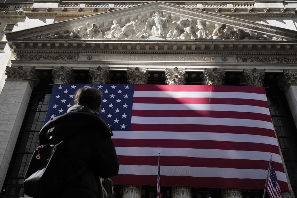 Mulher olha para bandeira dos Estados Unidos pendura em prédio da Bolsa de Valores de Nova York, em 26 de fevereiro de 2025. — Foto: Seth Wenig/AP