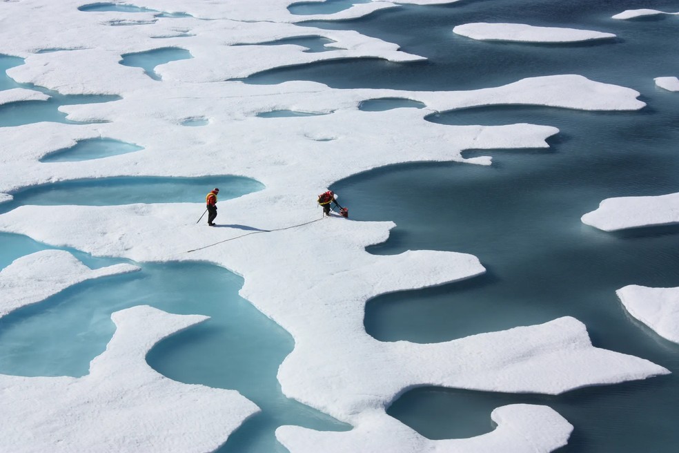 Duas pessoas caminham sobre placas de gelo, com algumas áreas derretidas em uma região do Ártico. — Foto: NASA/Kathryn Hansen