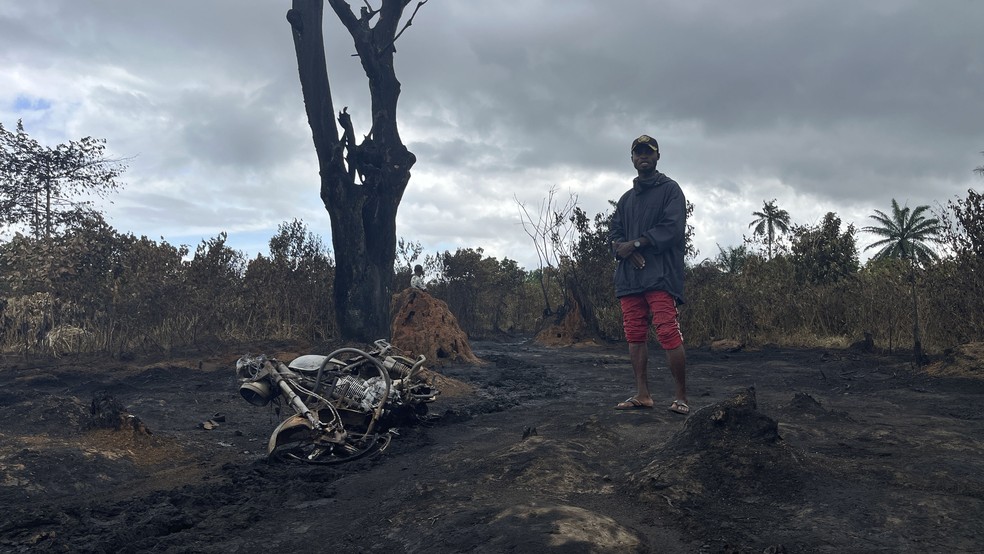 Homem fica de pé ao lado de uma árvore e uma motocicleta atingidos pela explosão em uma refinaria ilegal em Emohua, na Nigéria, em 3 de outubro de 2023 — Foto: Associated Press