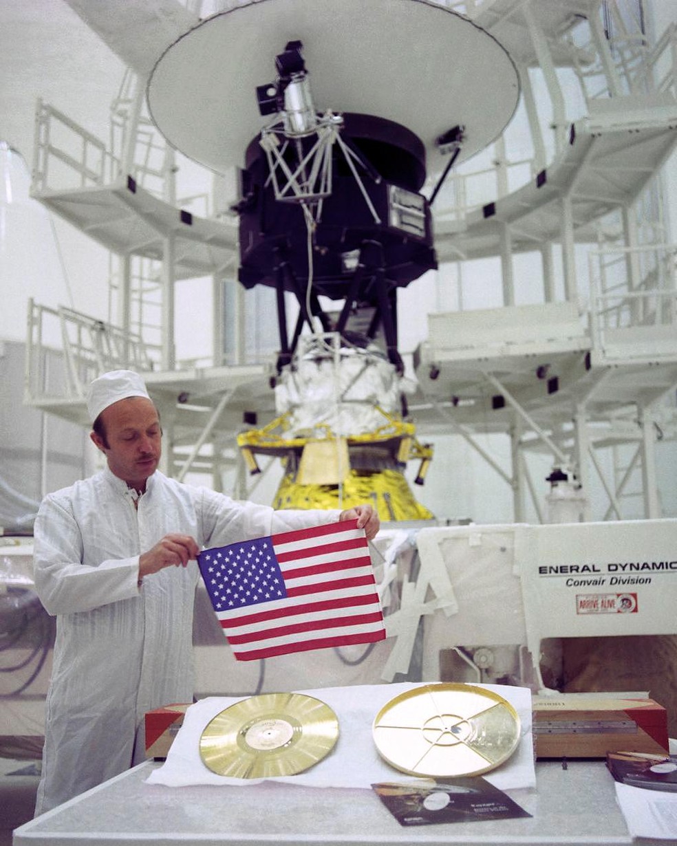 John Casani, gerente de projeto da Voyager, segura uma bandeira dos EUA que foi acoplada na Voyager 2. As duas sondas também levam discos fonográficos com imagens que retratam a vida na Terra. — Foto: Nasa