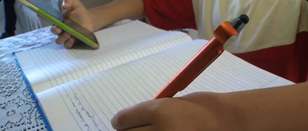 Imagem ilustrativa mostra mãos de aluno segurando celular e caneta — Foto: Reprodução/TV Gazeta