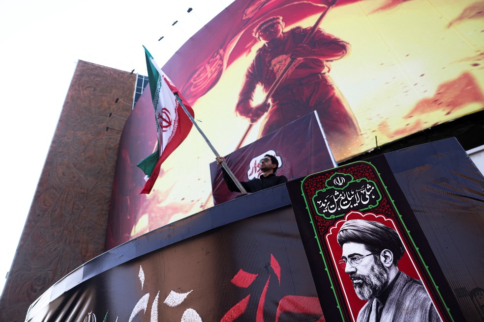 Homem segura bandeira do Irã perto de foto do líder supremo, Mojtaba Khamenei, em Teerã, em 13 de agosto de 2026. — Foto: Majid Asgaripour/WANA via Reuters
