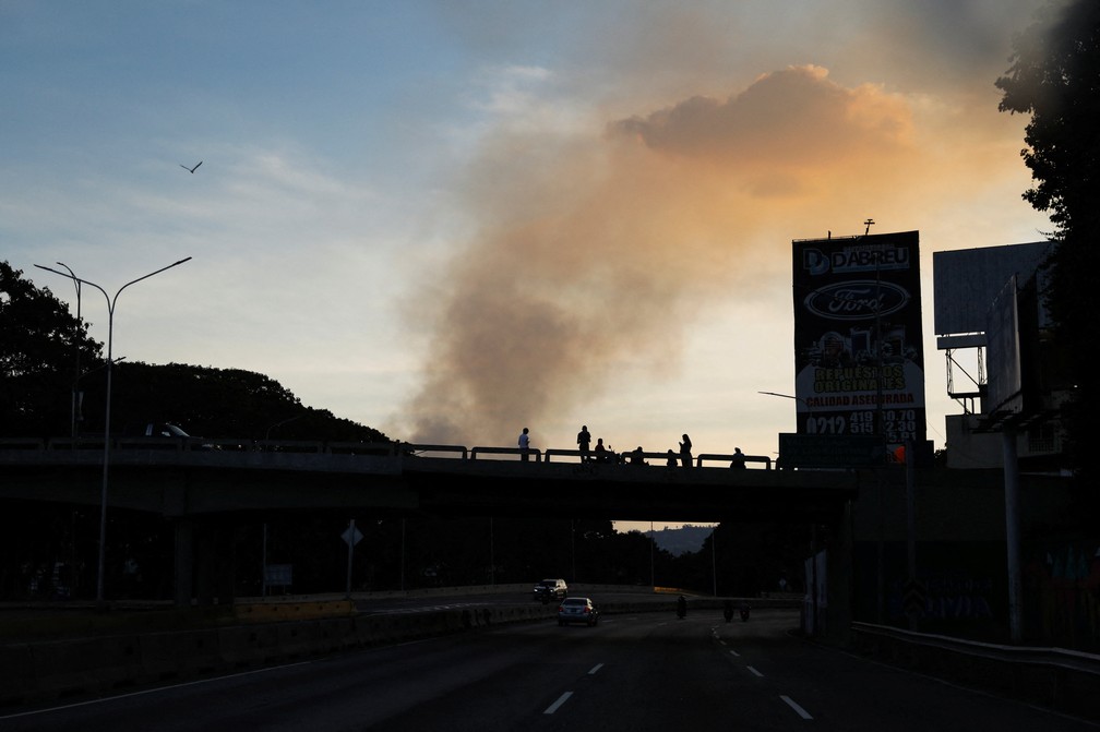 Pessoas em uma ponte são vistas em silhueta enquanto a fumaça sobe perto do Forte Tiuna após ataques dos EUA à Venezuela. — Foto: REUTERS/Leonardo Fernandez Viloria