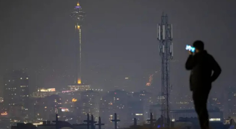 Um homem em Teerã usa o celular em uma colina com vista para a torre de telecomunicações Milad (foto de arquivo de 2024) — Foto: BBC/NurPhoto / Getty Images