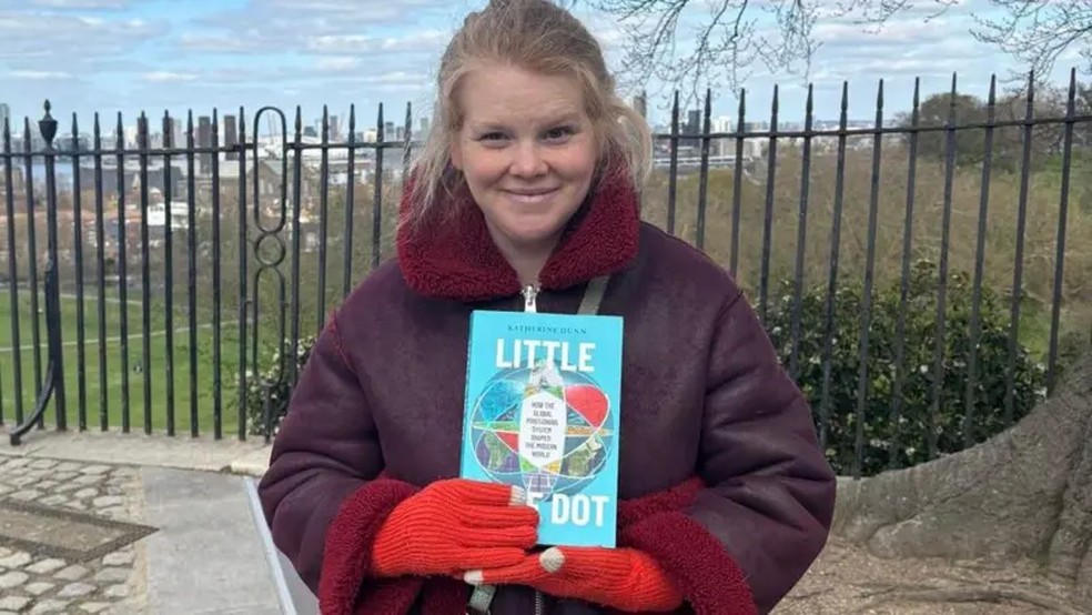 Katherine Dunn, no meridiano de Greenwich, com seu livro — Foto: Acervo Pessoal via BBC