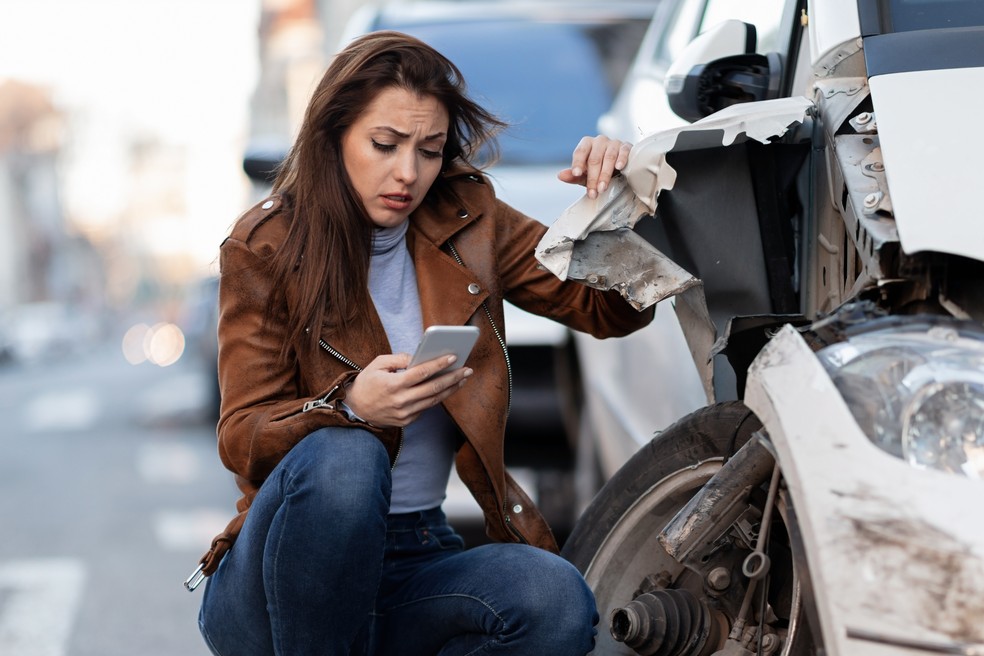 Mulher agachada com o celular na mão ao lado do carro batido  — Foto: Reprodução